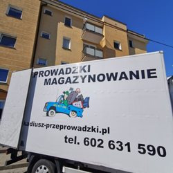 Biała furgonetka z napisem 'PROWADZKI MAGAZYNOWANIE', ilustracją niebieskiego samochodu z bagażami na dachu i adresem strony internetowej oraz numerem telefonu, zaparkowana przed żółtym blokiem...