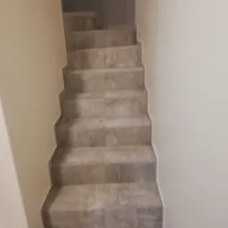 Wyłożone szarymi płytkami schody wewnętrzne prowadzące w górę, z widocznym fragmentem pomalowanej na beżowo ściany po prawej stronie i ciemnym przejściem u szczytu schodów.