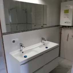 Nowoczesna, jasna łazienka z białą umywalką z szafką, dużym lustrem odbijającym kabinę prysznicową i grzejnik, oraz wiszącym piecem grzewczym.