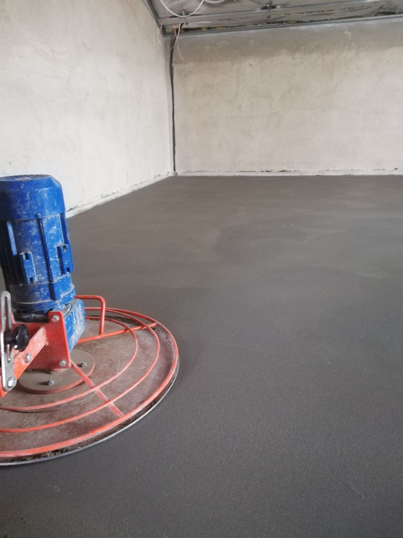 Zacieraczka mechaniczna do betonu podczas pracy na świeżo wylanym, szarym podkładzie podłogowym w surowym pomieszczeniu z widocznymi instalacjami pod sufitem.