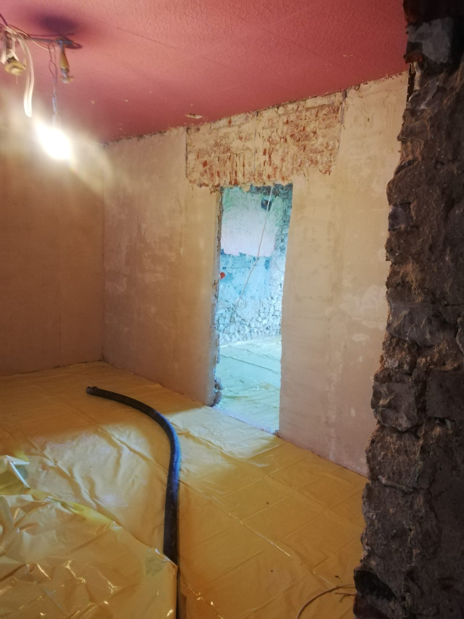 Przygotowanie pomieszczenia do wylewki: Folia ochronna na podłodze, ściany częściowo otynkowane, widoczny wąż do pompy, sufit w kolorze różowym.