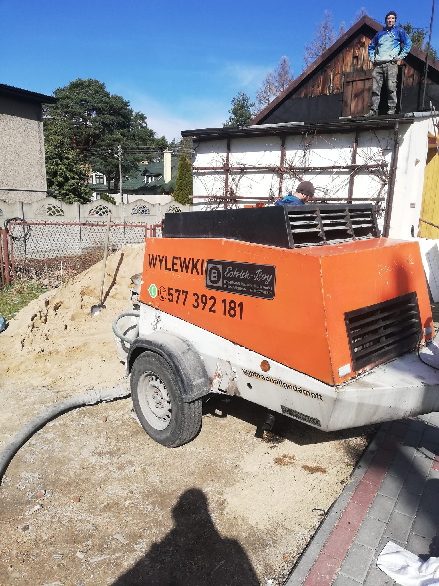 Pomarańczowo-biała maszyna do wylewek z widocznym logo i numerem telefonu, obok pryzma piasku i łopata, w tle pracownik na dachu i budynki mieszkalne.