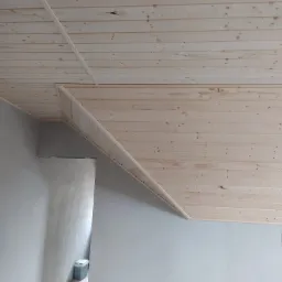 Drewniana boazeria na skośnym suficie poddasza, widok na fragment ściany pomalowanej na szaro, w tle widoczny fragment podłogi i pojemnik.