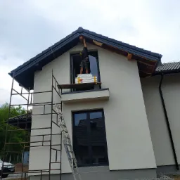 Pracownik na rusztowaniu przy oknie domu, przygotowujący się do montażu elementów wykończeniowych pod dachem, z widoczną elewacją w neutralnym odcieniu szarości i fragmentem dachu z ciemną dachówką.