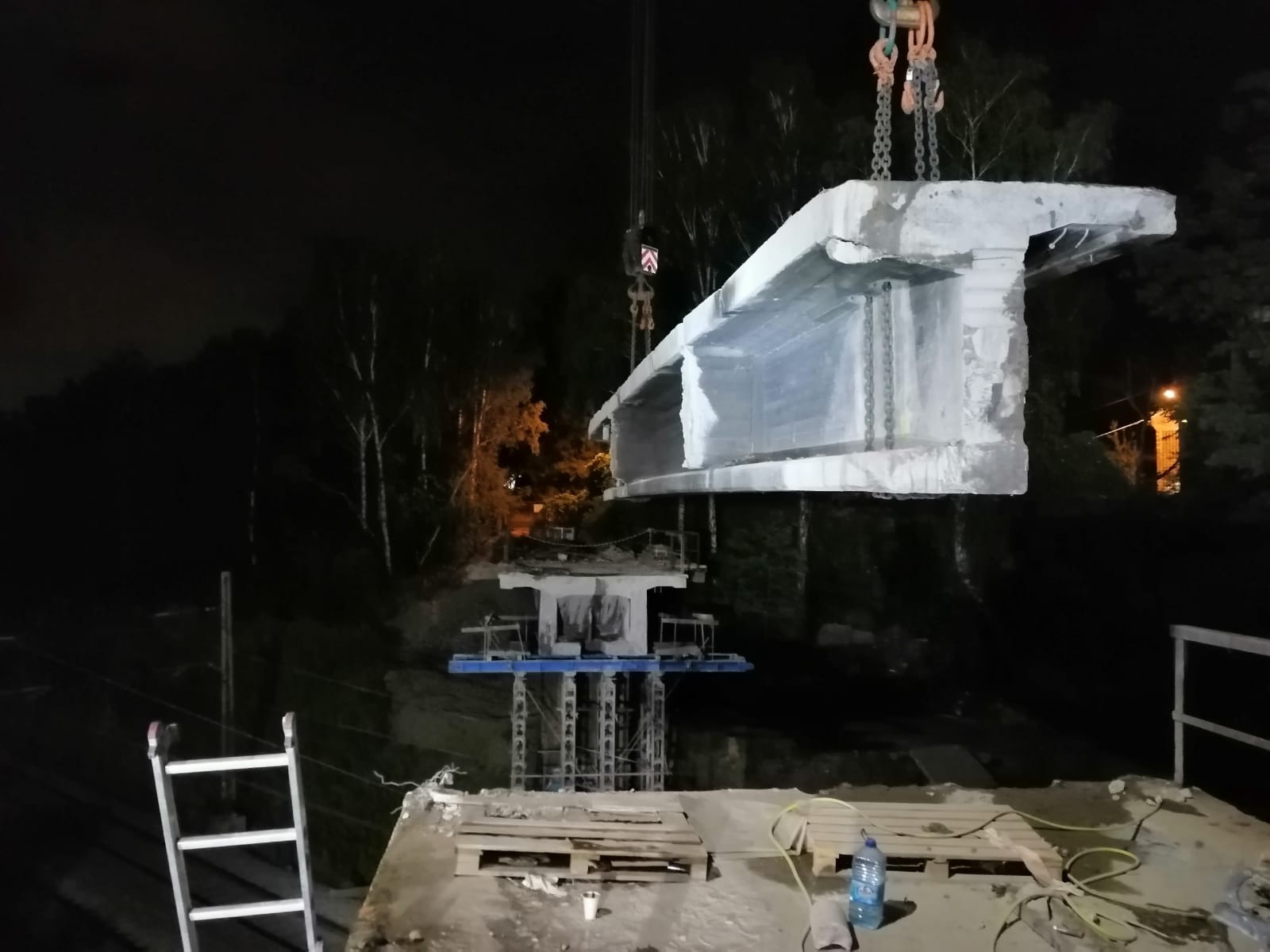 Montaż betonowego elementu mostu dźwigiem na tle nocnego nieba i drzew, widoczna drabina i butelka wody.