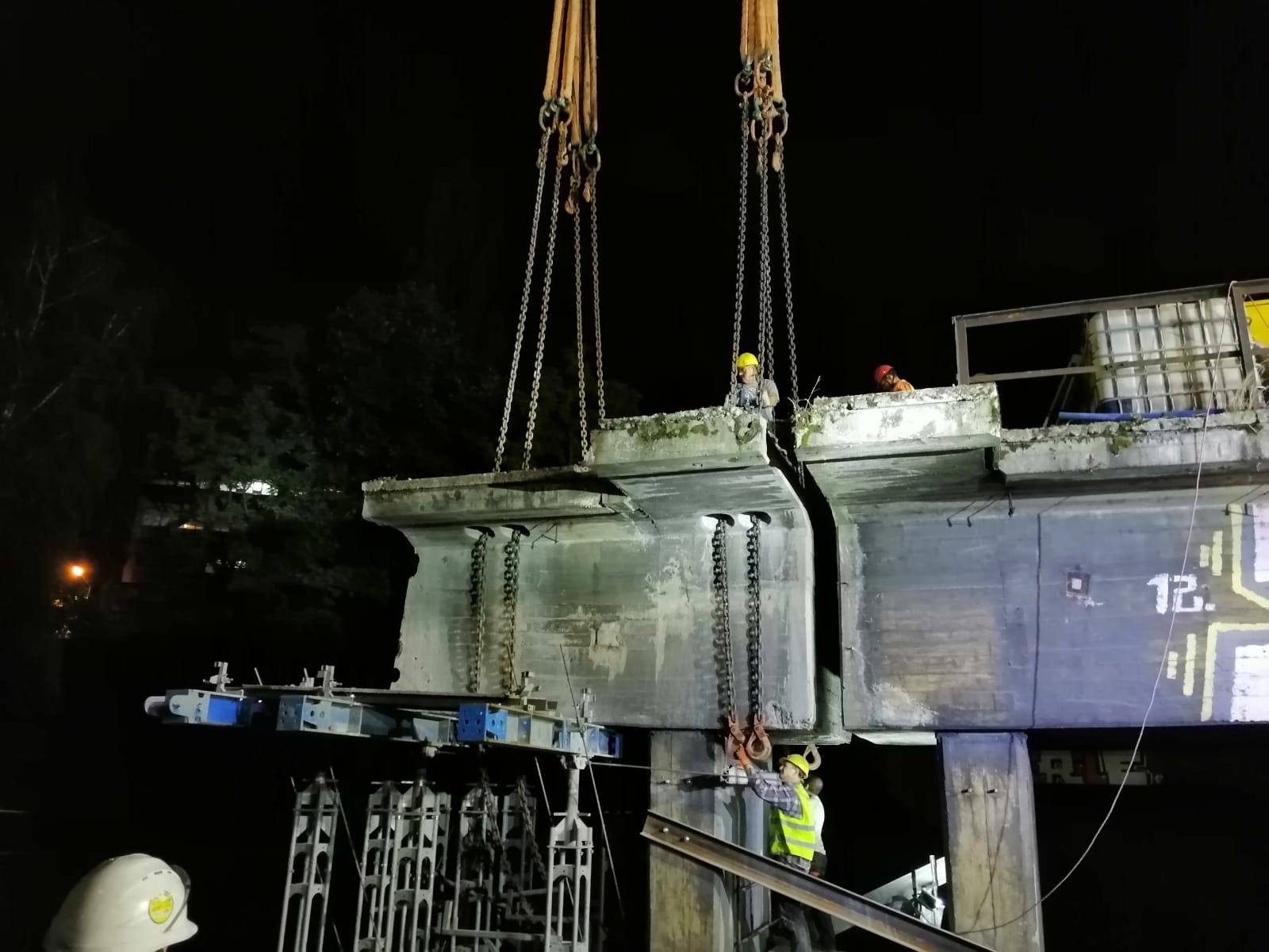 Demontaż przęsła mostu nocą, pracownicy w kaskach i kamizelkach odblaskowych, dźwig z łańcuchami, fragment konstrukcji z numerem '12'.