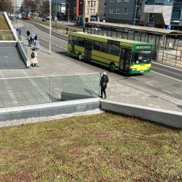 OK Projekt Jakub Okularczyk - Zielony dach budynku w miejskiej scenerii z autobusem w tle. Widok z góry, podkreślający roślinność na dachu i architekturę otoczenia.
