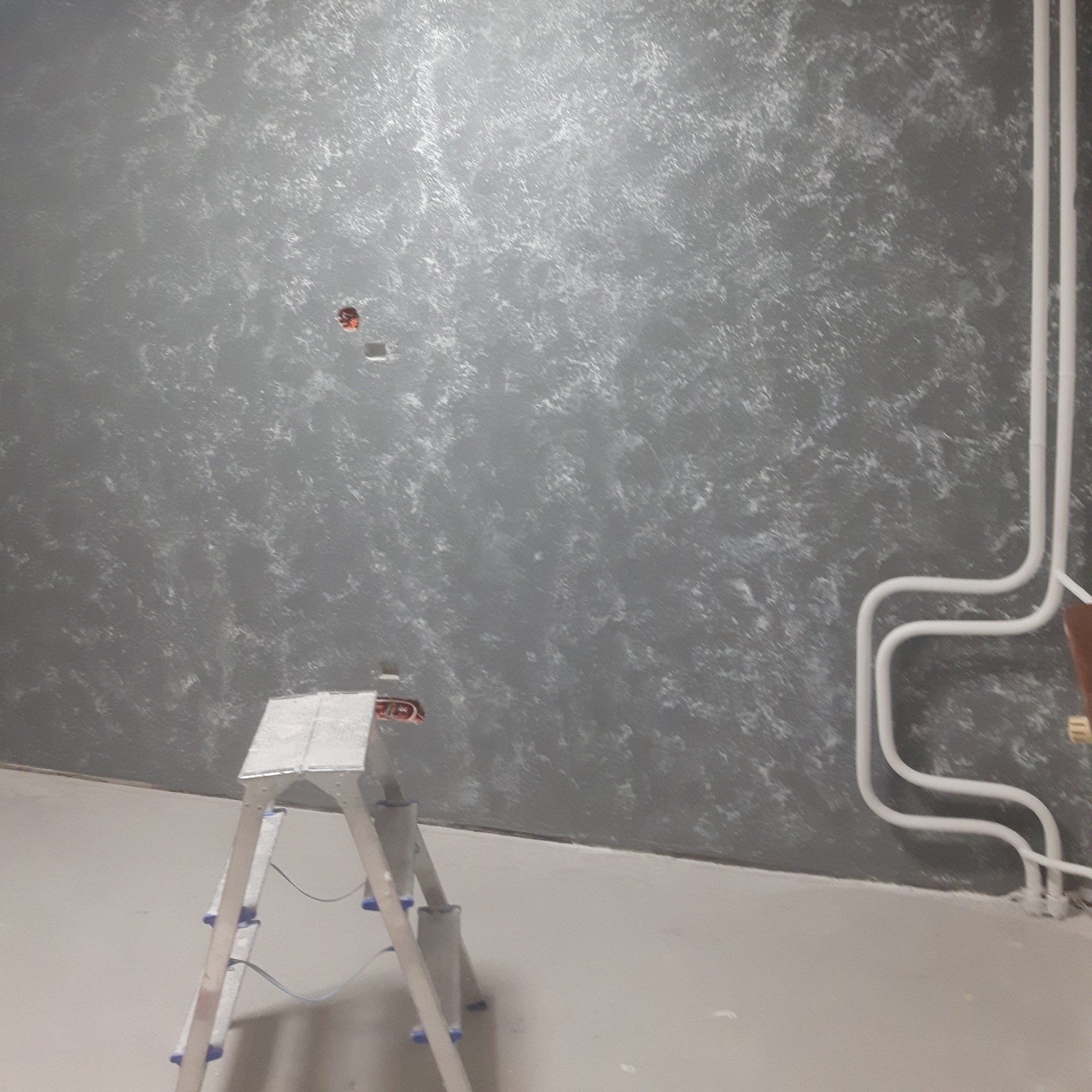 Ściana z efektem dekoracyjnym w odcieniach szarości, widoczna drabina malarska i instalacja rur przy podłodze.