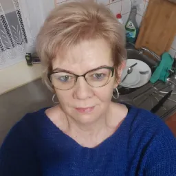 Portret kobiety w okularach, w tle zlewozmywak z naczyniami i środki czystości, sugerujący domowe otoczenie.