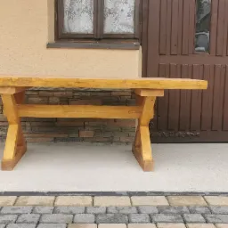 Drewniany stół ogrodowy z krzyżowymi nogami, ustawiony na betonowej posadzce obok ciemnego samochodu i brązowych drzwi. Na samochodzie naklejka z napisem 'Don't touch my Alfa Romeo'.