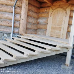 Surowe drewniane schody prowadzące do wejścia domu z bali, widoczne detale zdobień na drzwiach i drewnianej elewacji, szara rynna i kamienna podmurówka.