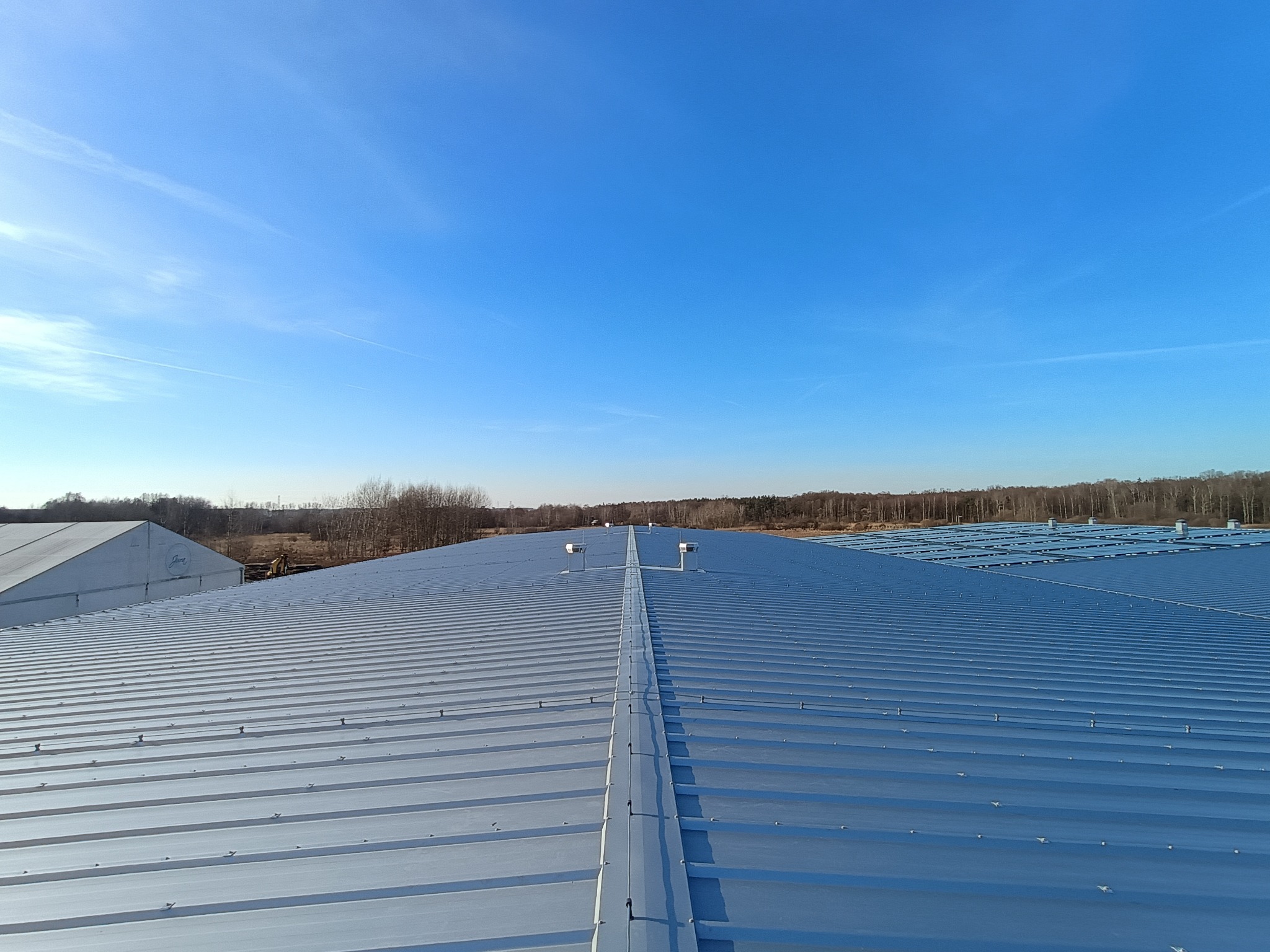 Widok z góry na rozległy dach hali przemysłowej pokryty blachą trapezową w kolorze srebrnym, z widocznymi elementami mocującymi i systemem wentylacji, na tle błękitnego nieba i zalesionego terenu.