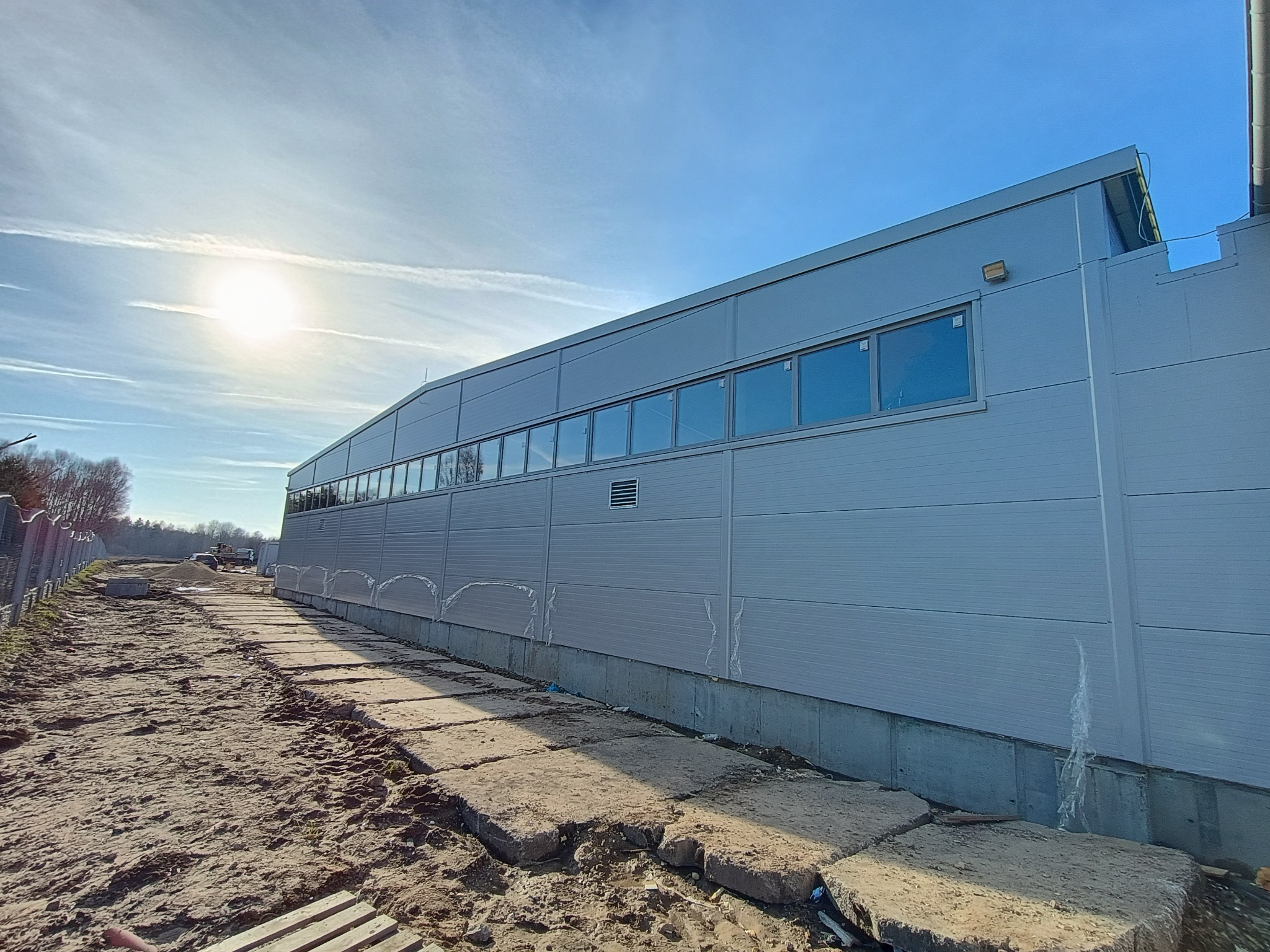 Długa hala przemysłowa z szarej płyty warstwowej, widok z boku, z rzędem okien i betonową podstawą. Teren wokół w stanie surowym, z widocznymi śladami opon i resztkami folii budowlanej przy...