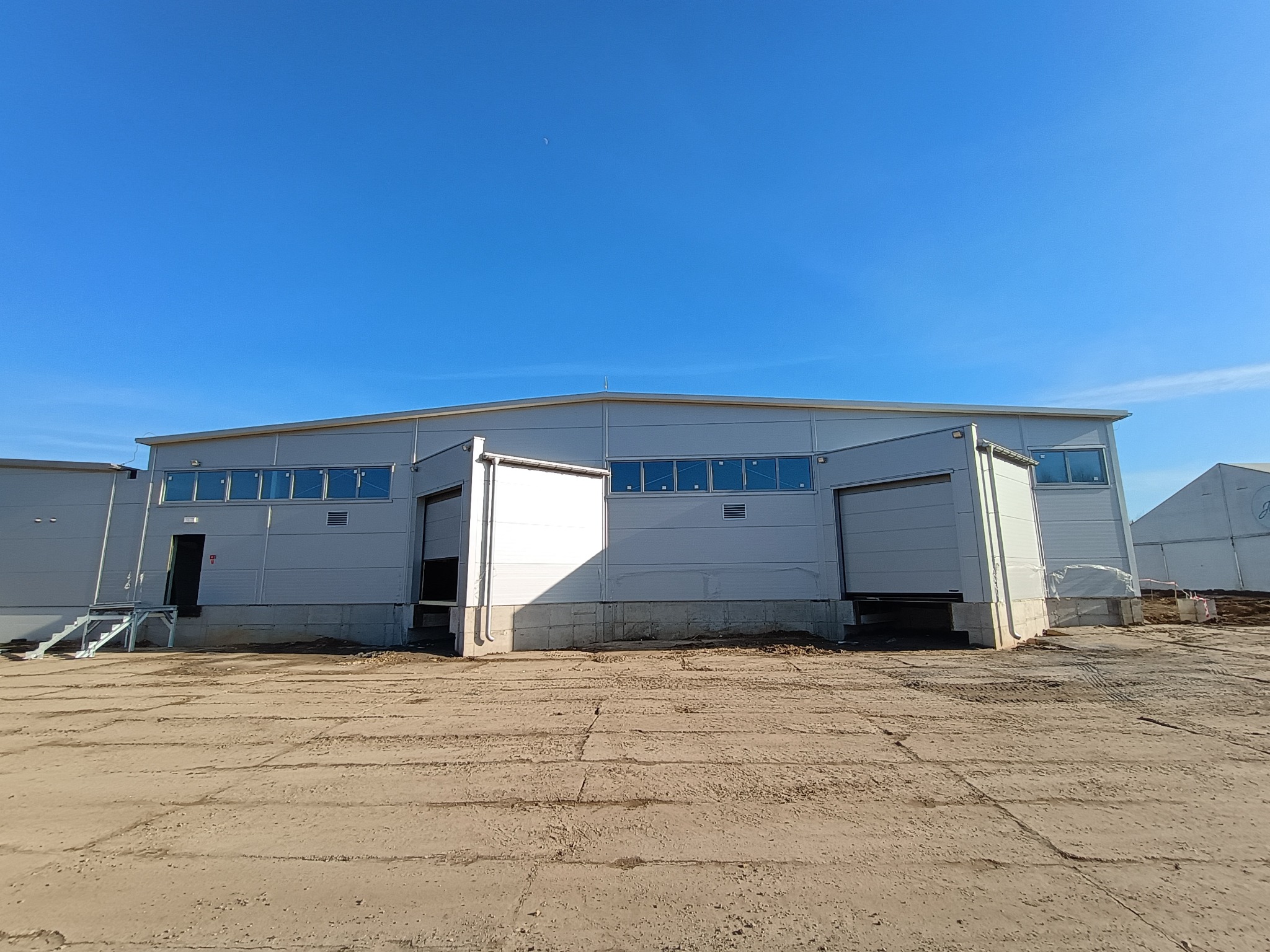 Nowoczesna hala magazynowa z szarej płyty warstwowej, z otwartymi bramami rolowanymi i schodami wejściowymi, na betonowym placu w słoneczny dzień.