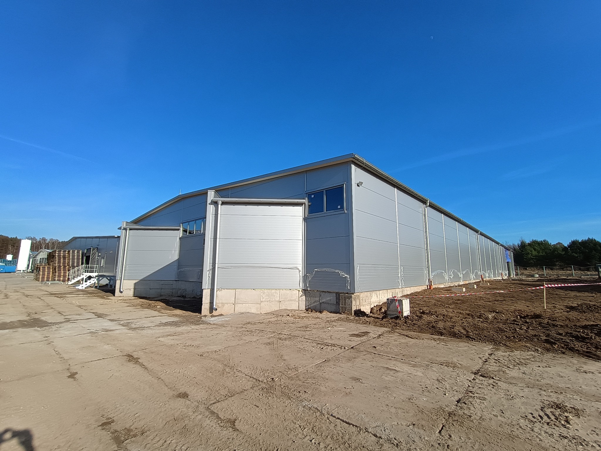 Nowo wybudowana hala magazynowa z jasnoszarych płyt warstwowych, widok z zewnątrz, teren wokół hali częściowo utwardzony, częściowo z ziemią i wyznaczonym terenem budowy.