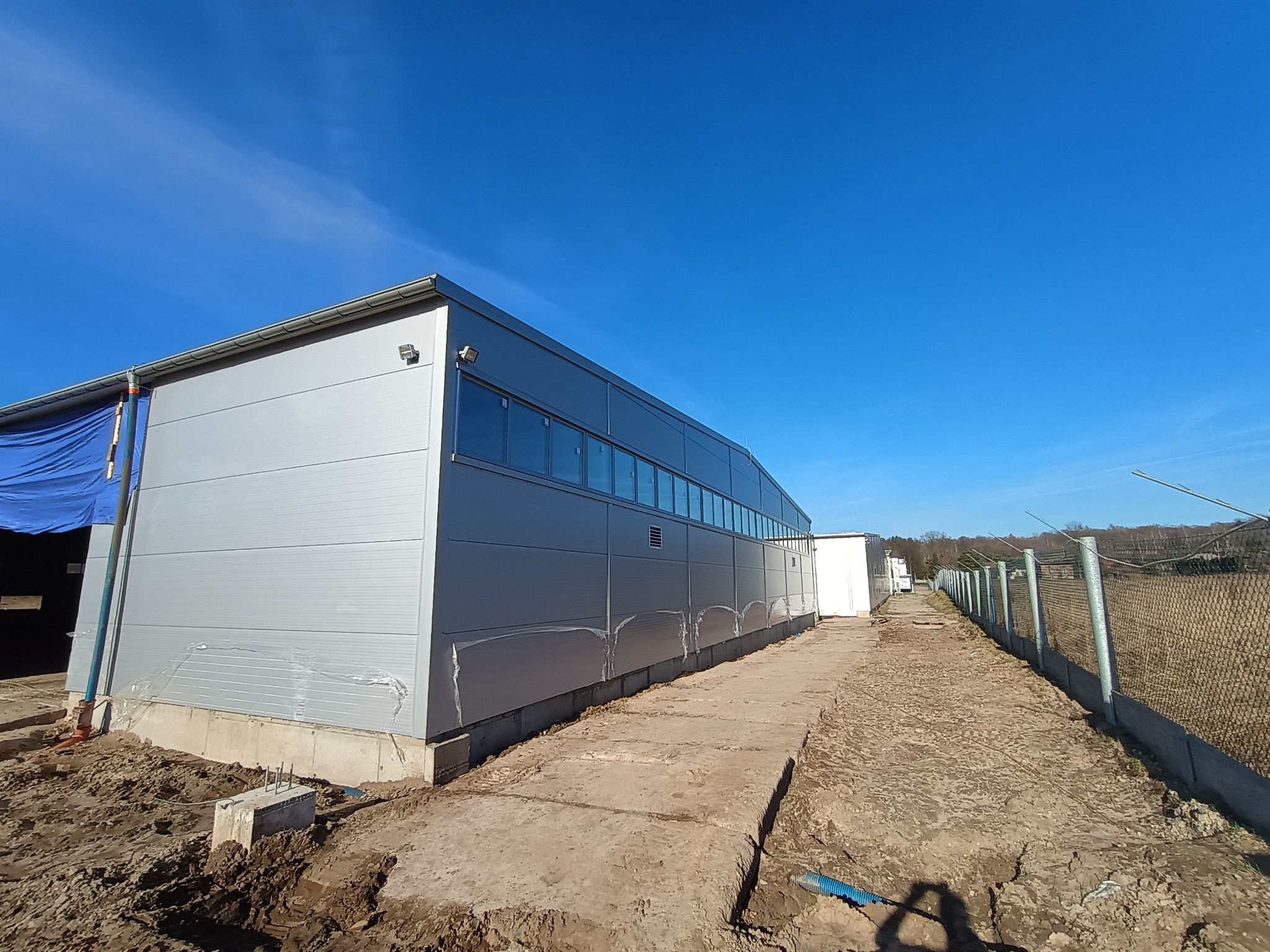 Hala przemysłowa w trakcie budowy, widoczne płyty warstwowe na elewacji, fundamenty i teren wokół w stanie surowym, błękitne niebo w tle.