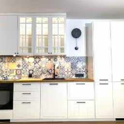 Biała zabudowa kuchenna z drewnianym blatem i kolorową mozaiką na ścianie, uzupełniona czarną armaturą i wiszącym zegarem.