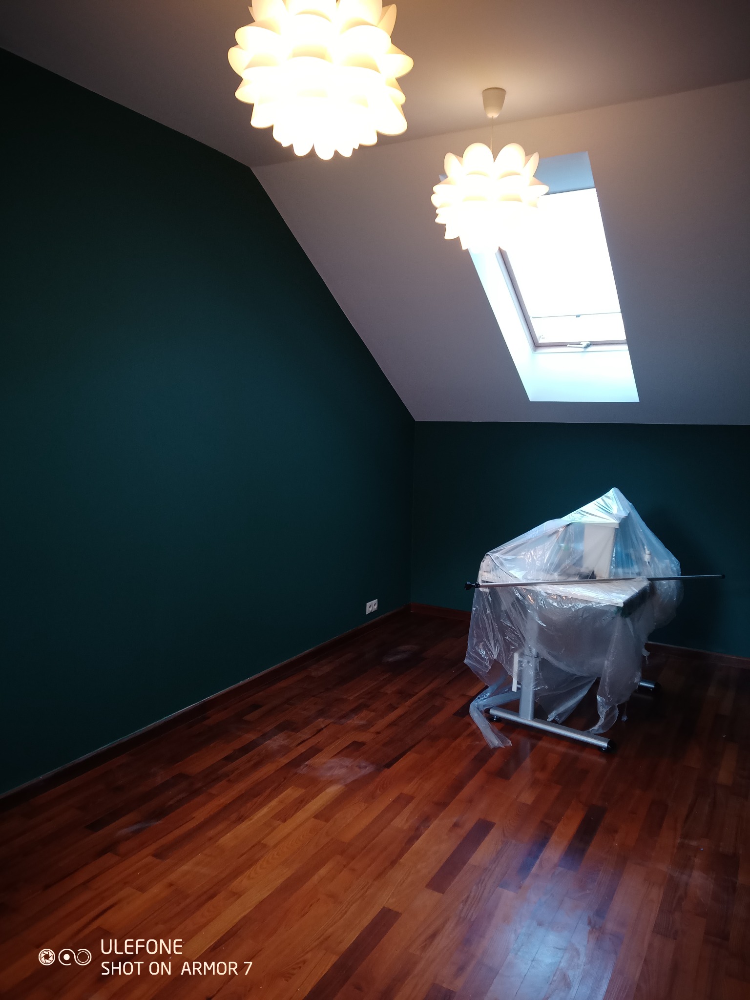 Pokój na poddaszu z ciemnozielonymi ścianami, drewnianą podłogą i oknem dachowym; meble zabezpieczone folią malarską przed zabrudzeniem.