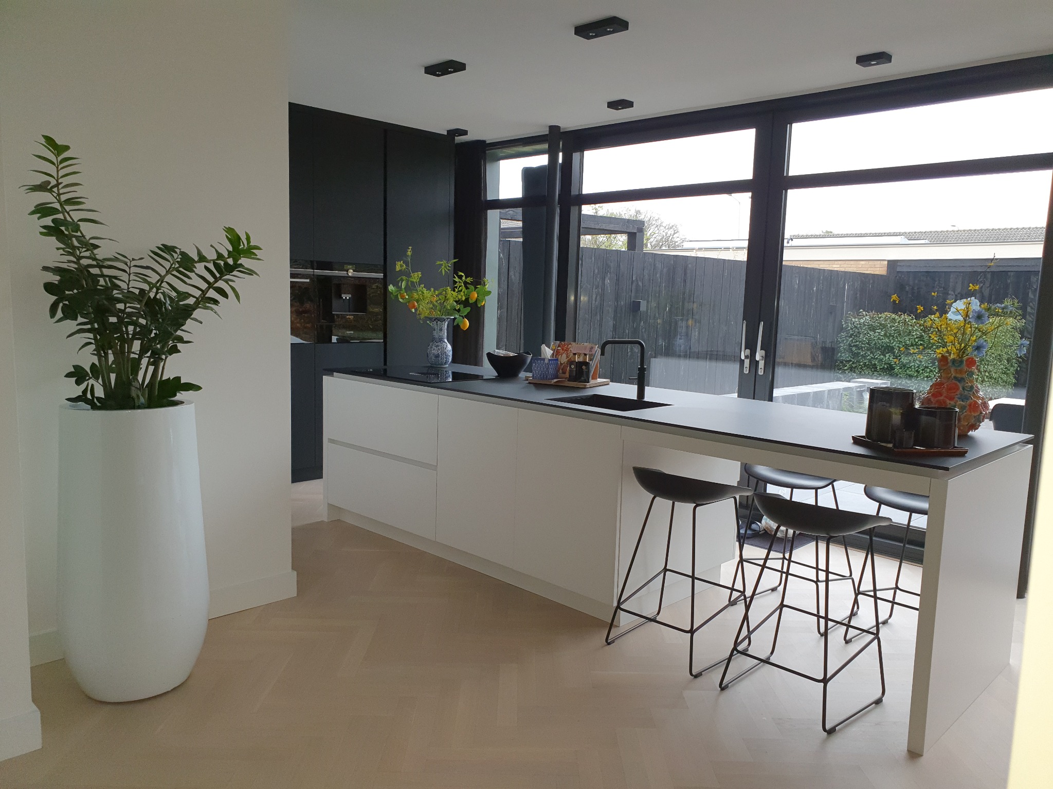 Minimalistyczna, jasna kuchnia z wyspą, czarnymi szafkami i dużym oknem z widokiem na zadbany ogród, uzupełniona dekoracjami kwiatowymi i nowoczesnymi stołkami barowymi.