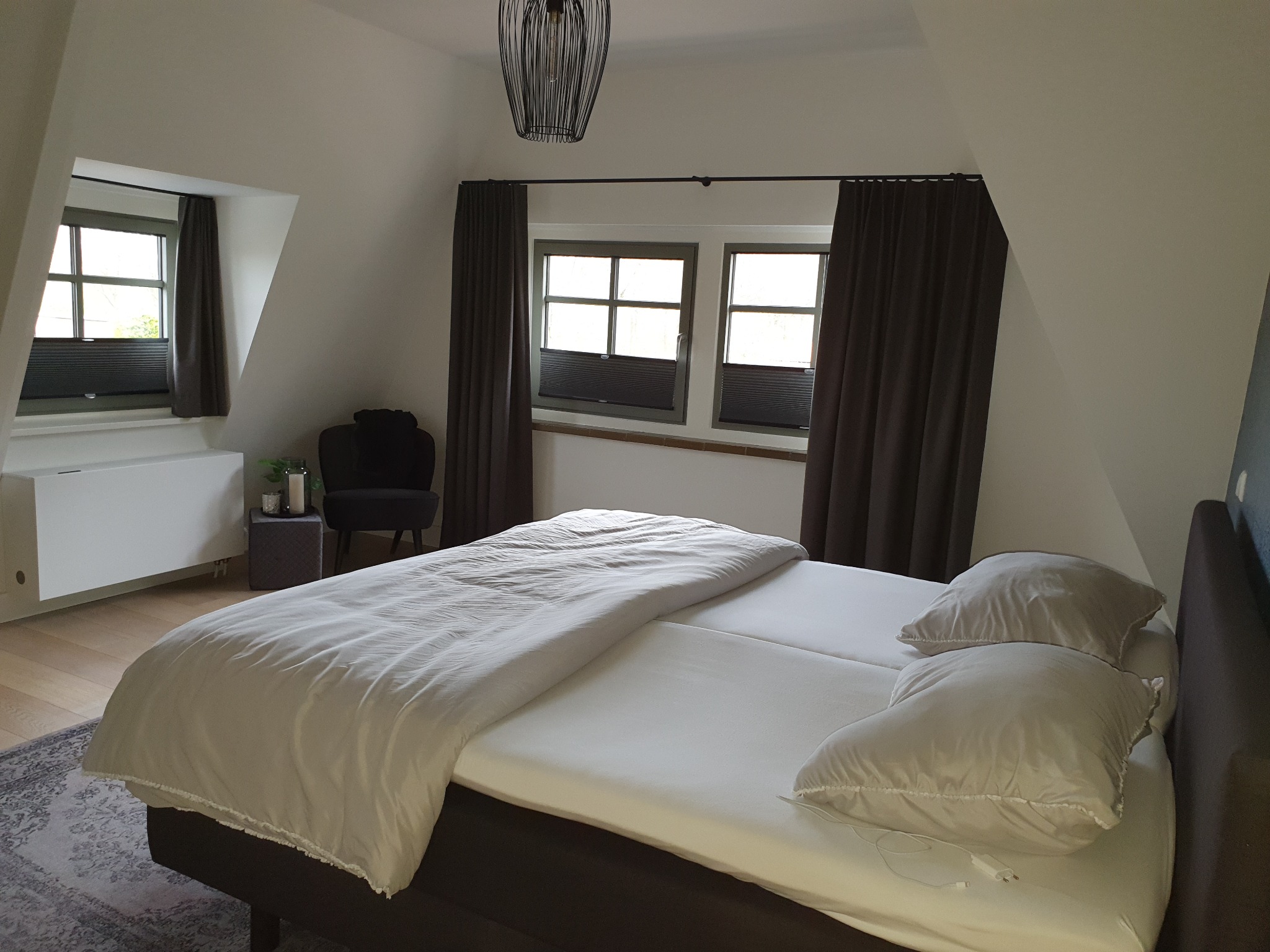 Jasna sypialnia na poddaszu z łóżkiem, oknami z roletami i ciemnymi zasłonami, widok na lampę wiszącą i dywan w tle.