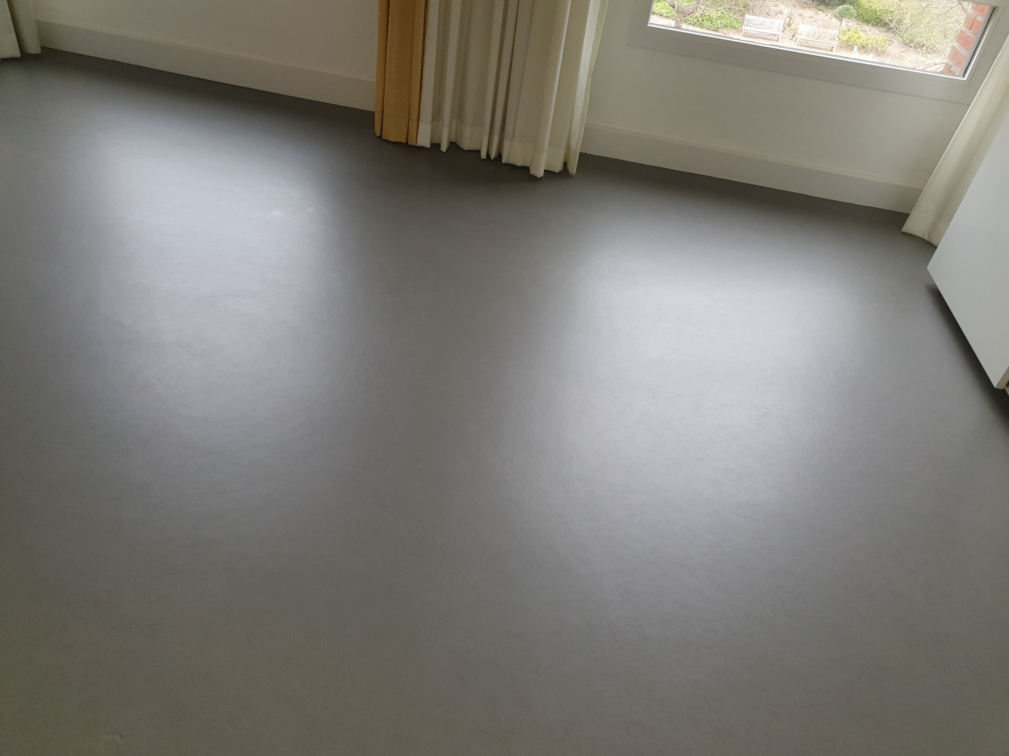 Gładka, szara podłoga w pustym pokoju z oknem i jasnymi zasłonami, widok z góry na świeżo umytą powierzchnię.