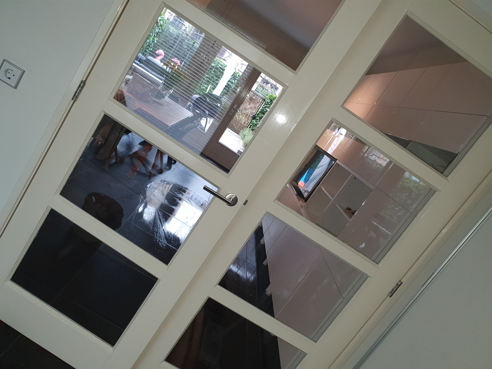 Białe, przeszklone drzwi wewnętrzne z widokiem na czarną podłogę, jasne płytki i elementy wyposażenia domu oraz ogród za oknem.