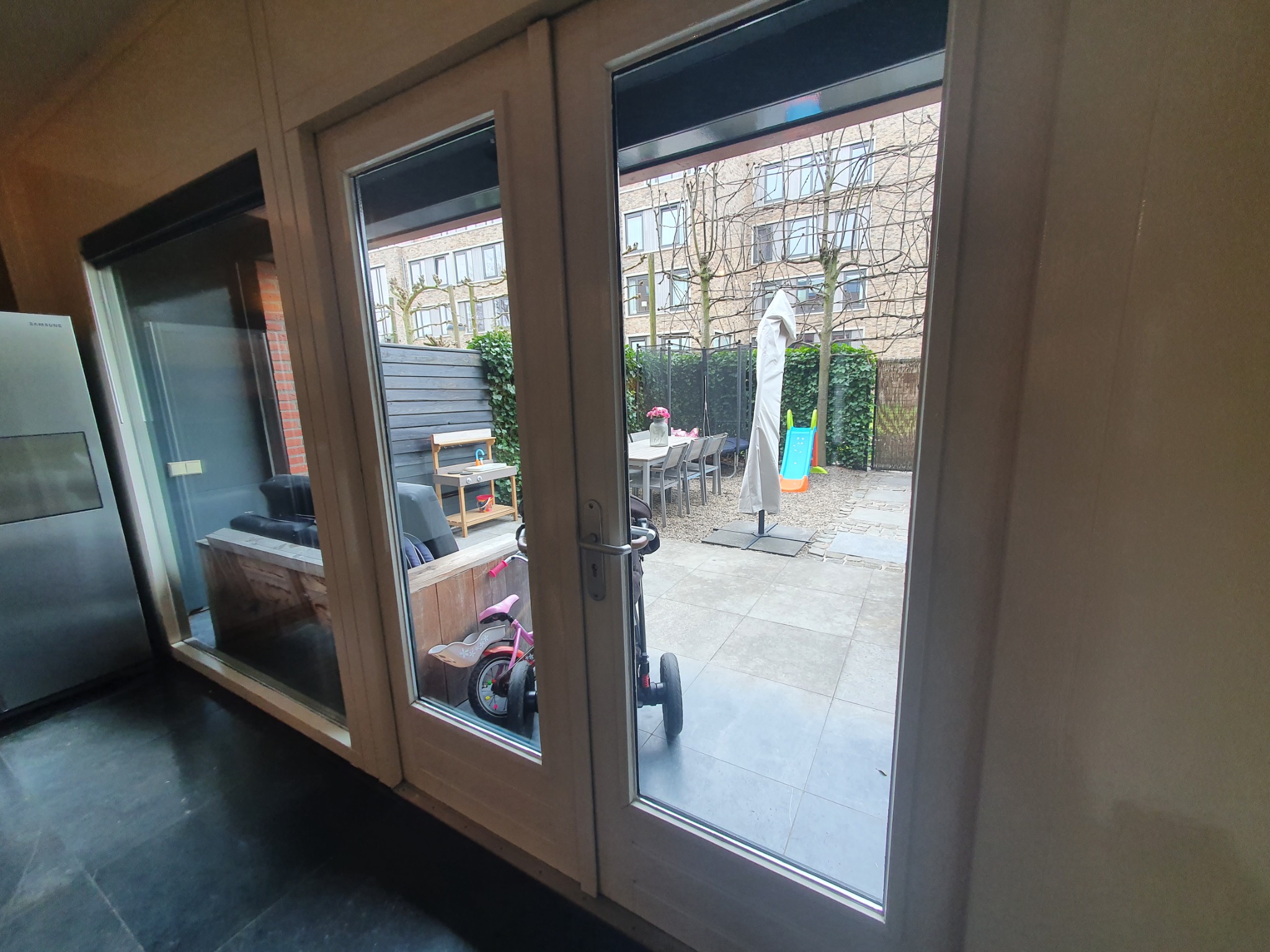 Widok przez przeszklone drzwi na zadbany ogród z meblami ogrodowymi, parasolem, trampoliną i zjeżdżalnią, z perspektywy wnętrza domu.