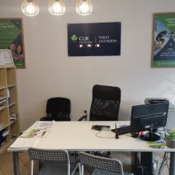 Wnętrze biura partnerskiego CUK Ubezpieczenia z biurkiem, komputerem, krzesłami, plakatem reklamowym ubezpieczeń turystycznych i logo firmy na ścianie.