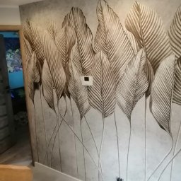 BIADACZ Paweł Biadacz - Ściana w pokoju z tapetą w motywie liści w odcieniach szarości i brązu, widoczny panel sterowania klimatyzacją i gniazdka elektryczne, fragment podłogi i widok na akwarium w sąsiednim pomieszczeniu.