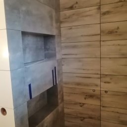 BIADACZ Paweł Biadacz - Ściana wyłożona płytkami imitującymi beton z wnękami w trakcie prac wykończeniowych, zabezpieczona niebieską taśmą, sąsiaduje z płytkami imitującymi drewno.