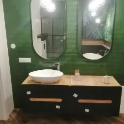 Nowoczesna łazienka z zielonymi ścianami, owalnymi lustrami i umywalką nablatową, widoczne niedbale naklejone naklejki na szafce umywalkowej.