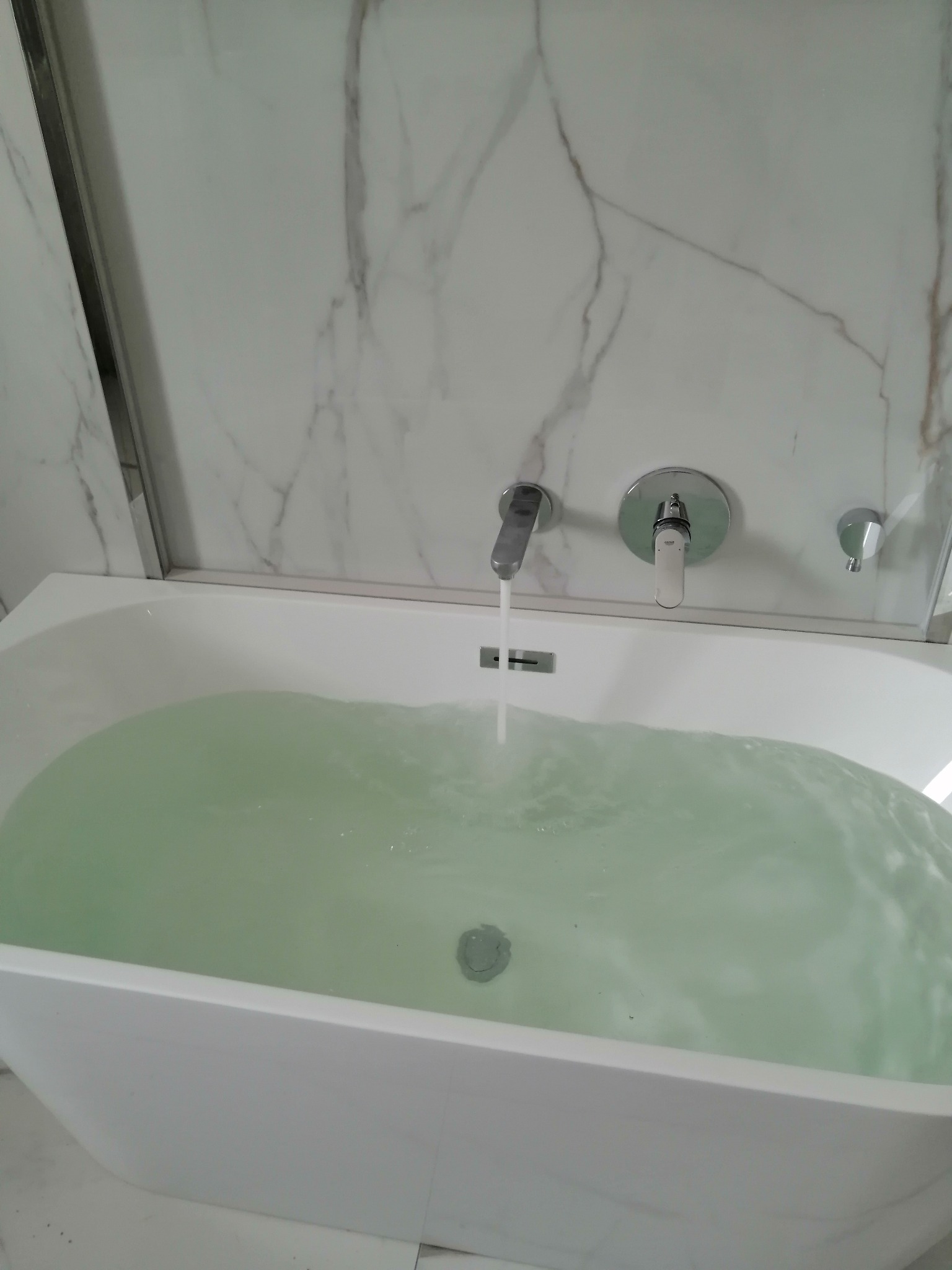 Woda nalewana do białej, owalnej wanny z chromowaną armaturą na tle marmurowej ściany z szklanym panelem.