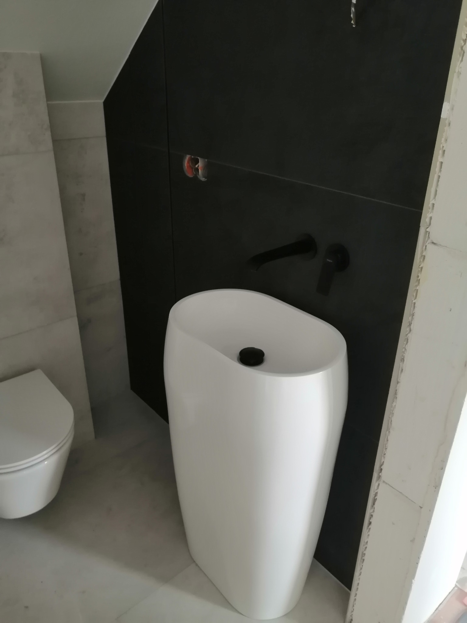 Nowoczesna łazienka z białą, wolnostojącą umywalką o opływowym kształcie i czarną armaturą na tle ciemnej ściany. Widoczny fragment toalety i jasna, marmurkowa podłoga.