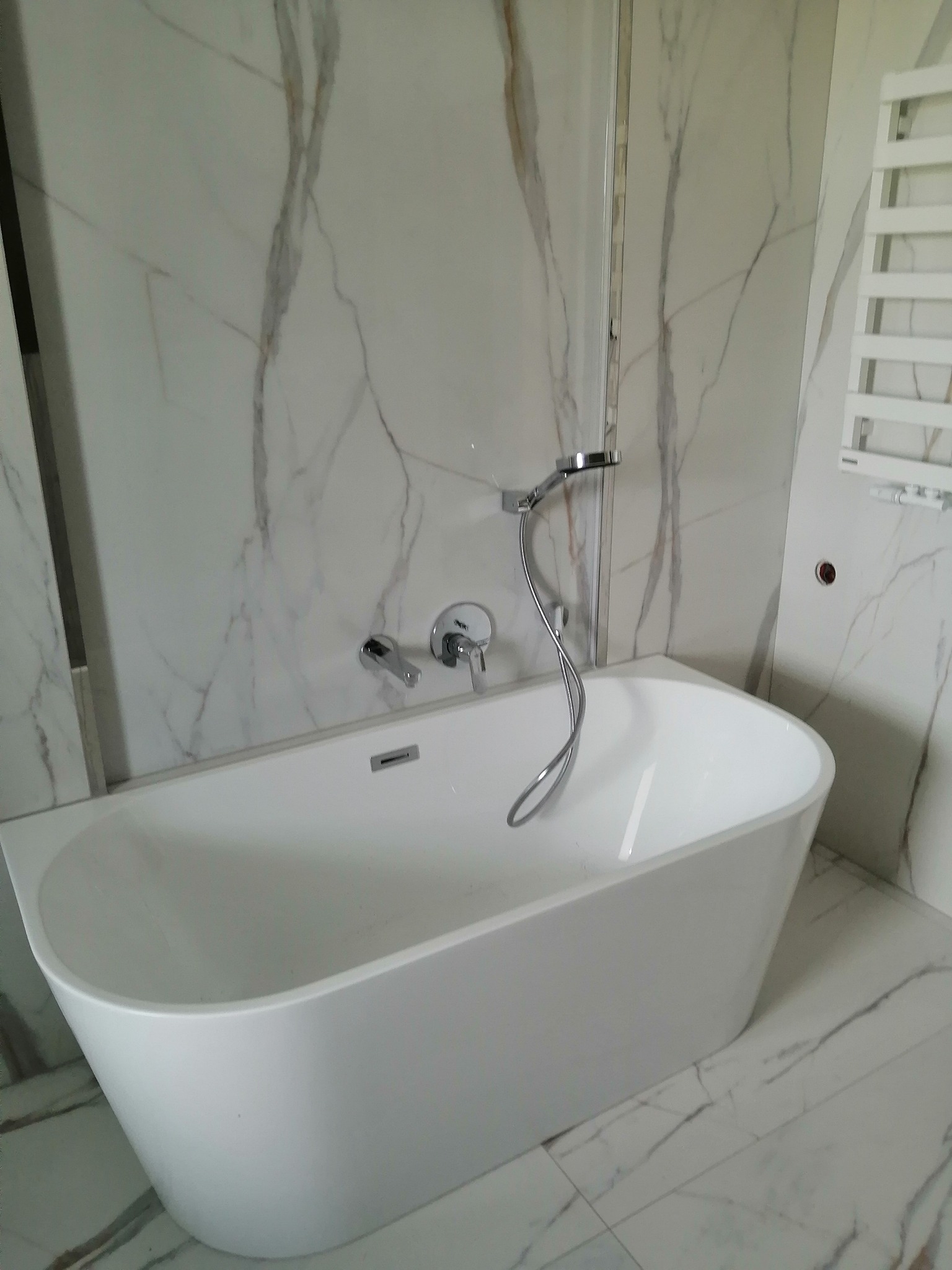 Nowoczesna biała wanna wolnostojąca z chromowaną armaturą i prysznicem, otoczona płytkami imitującymi biały marmur z szarymi żyłami, oraz drabinkowy grzejnik.