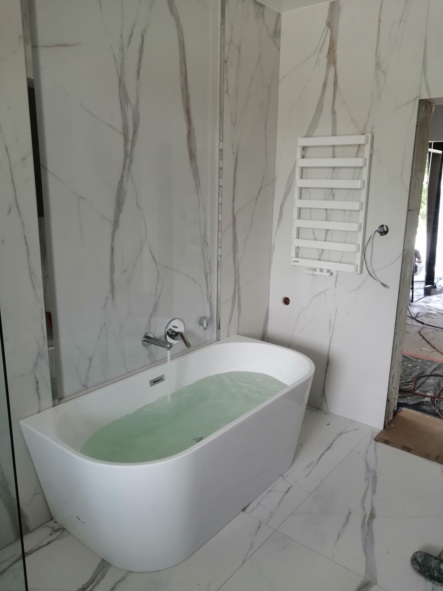 Nowoczesna, biała wanna wypełniona wodą, z chromowaną baterią. Ściany i podłoga wyłożone białymi płytkami imitującymi marmur. Widoczny biały grzejnik drabinkowy i fragment niezakończonych prac...