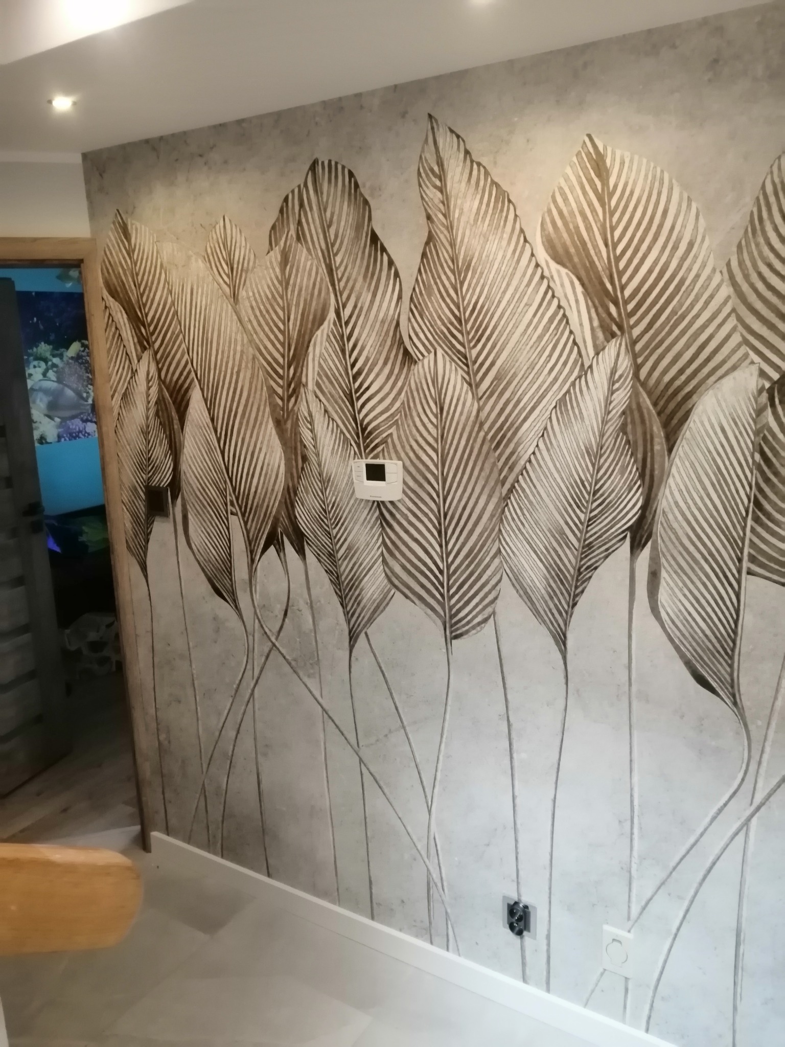 Ściana w pokoju z tapetą w motywie liści w odcieniach szarości i brązu, widoczny panel sterowania klimatyzacją i gniazdka elektryczne, fragment podłogi i widok na akwarium w sąsiednim pomieszczeniu.