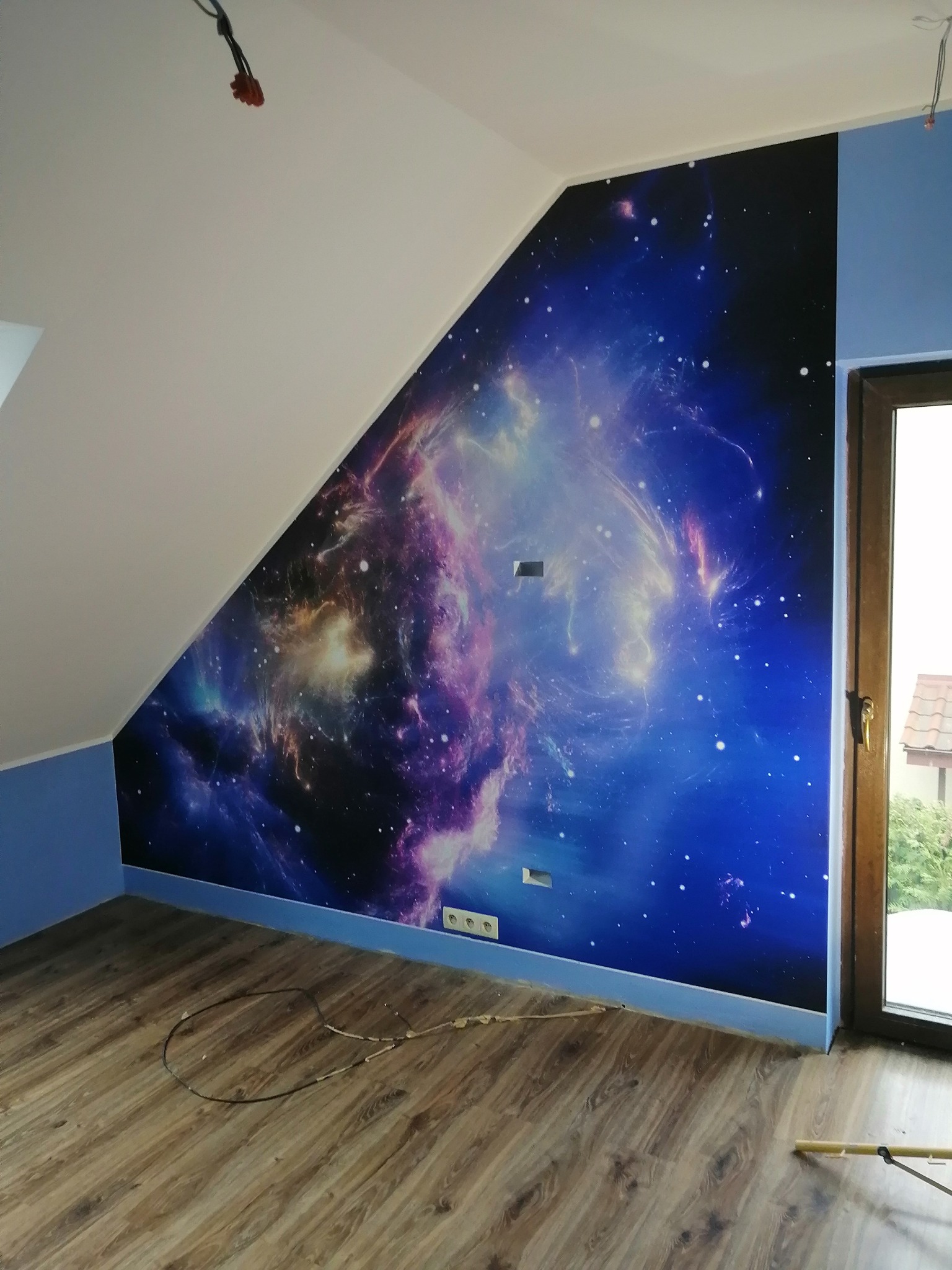 Pokój z fototapetą przedstawiającą kosmos na skośnej ścianie, podłoga z paneli, niebieskie wykończenie ścian, widoczne gniazdka elektryczne i przewody.