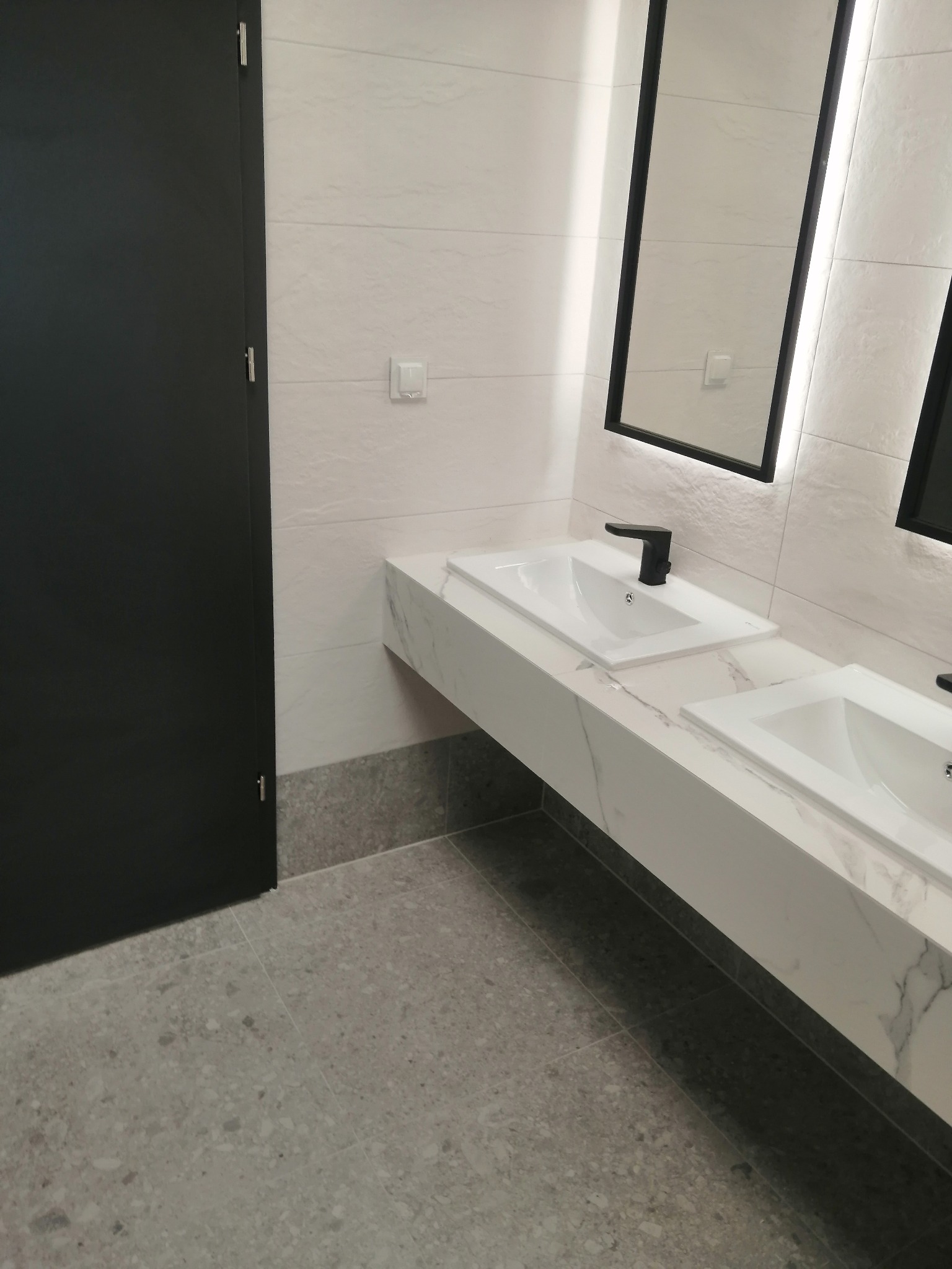 Nowoczesna łazienka z dwoma umywalkami nablatowymi na blacie imitującym marmur, czarną armaturą, podświetlanymi lustrami w czarnych ramach, jasnymi płytkami ściennymi i szarą podłogą z terazzo...