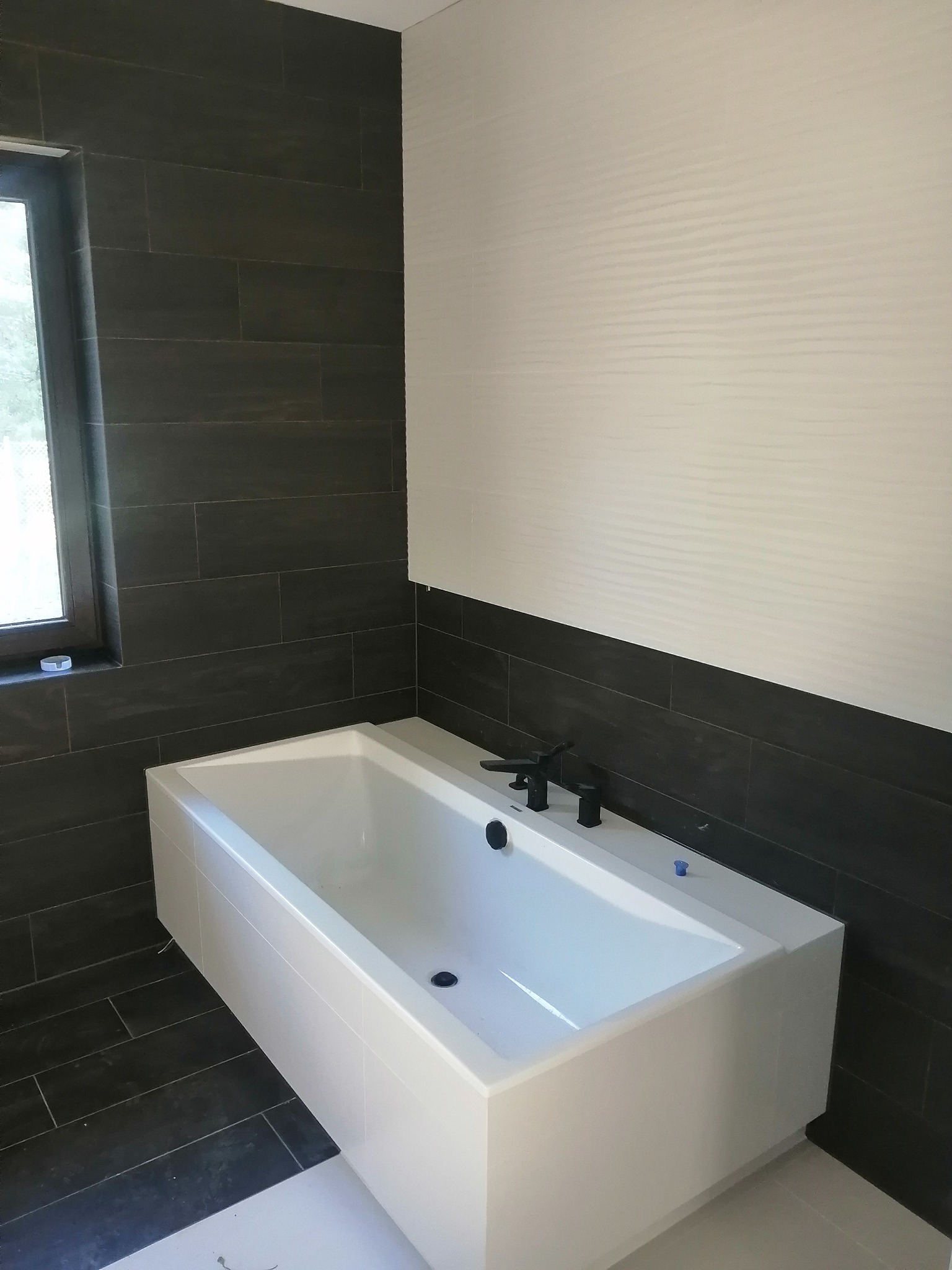 Nowoczesna, prostokątna wanna zabudowana płytkami w Aleksandrowie Kujawskim, z czarną armaturą na tle ściany wykończonej ciemnymi, podłużnymi płytkami i białymi, poziomymi panelami.