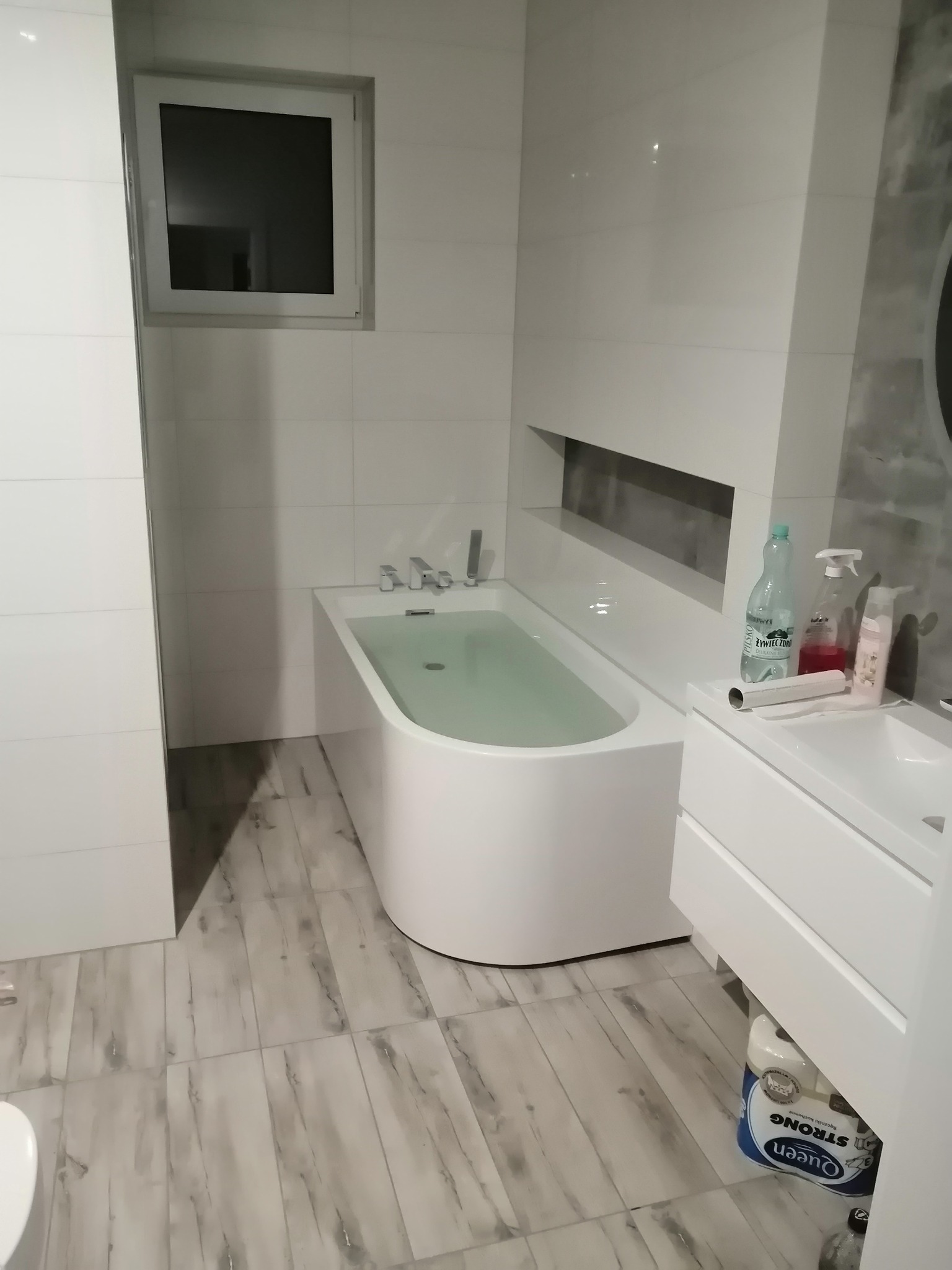 Nowoczesna, jasna łazienka z wolnostojącą wanną wypełnioną wodą, umywalką z białymi szafkami i szarą półką w ścianie. Na blacie umywalki stoją kosmetyki i butelka wody, a pod szafką leży papier...