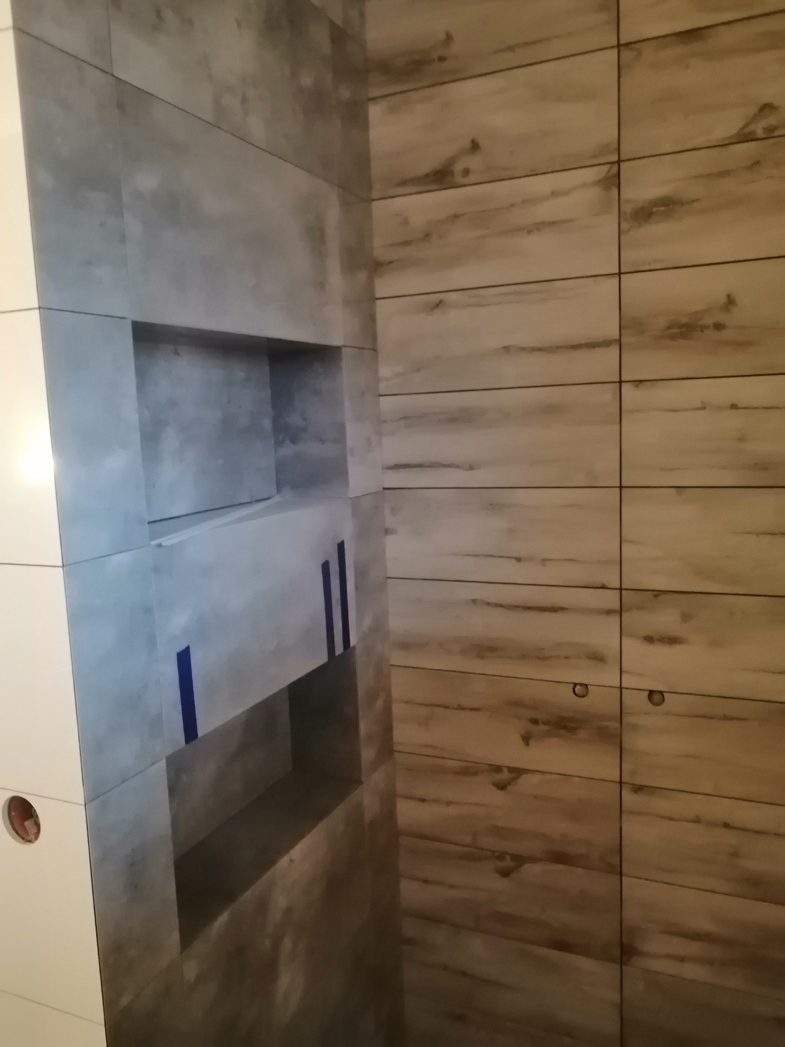 Ściana wyłożona płytkami imitującymi beton z wnękami w trakcie prac wykończeniowych, zabezpieczona niebieską taśmą, sąsiaduje z płytkami imitującymi drewno.