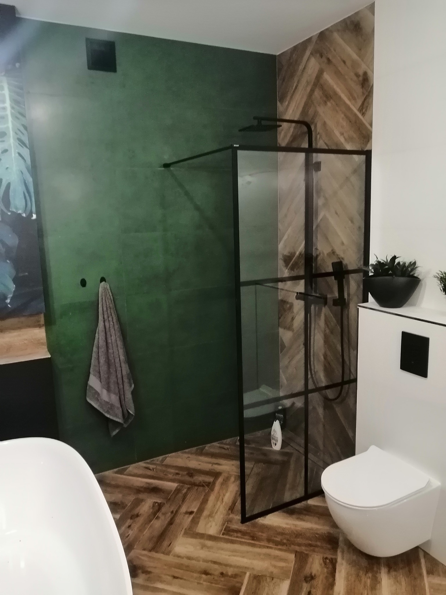 Nowoczesna łazienka z zieloną ścianą, kabiną prysznicową z czarnymi profilami, toaletą podwieszaną i podłogą w jodełkę z płytek imitujących drewno.