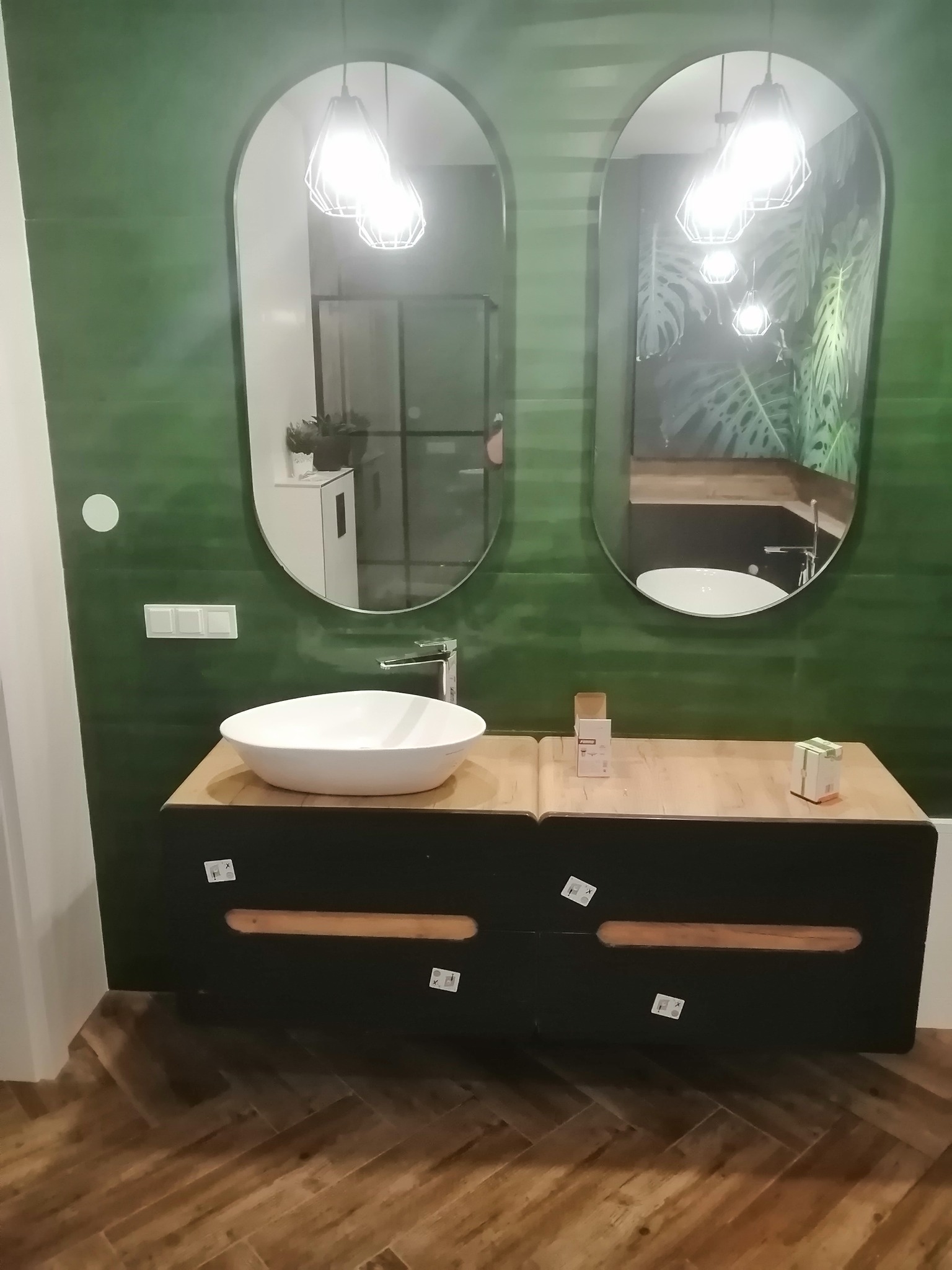 Nowoczesna łazienka z zielonymi ścianami, owalnymi lustrami i umywalką nablatową, widoczne niedbale naklejone naklejki na szafce umywalkowej.