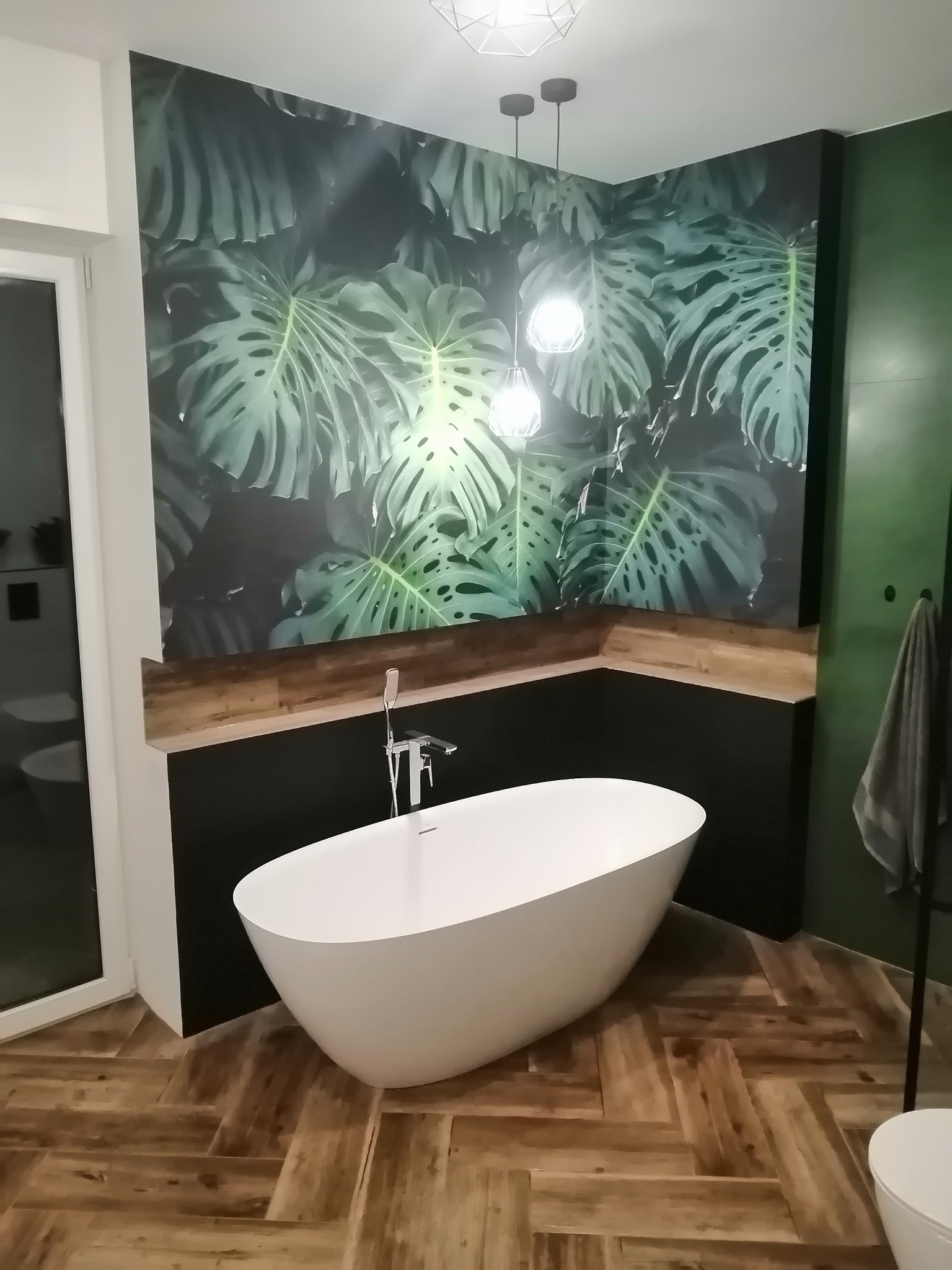 Nowoczesna łazienka z wolnostojącą wanną, drewnopodobną podłogą ułożoną w jodełkę, ścianą w tropikalne liście i wiszącymi lampami.