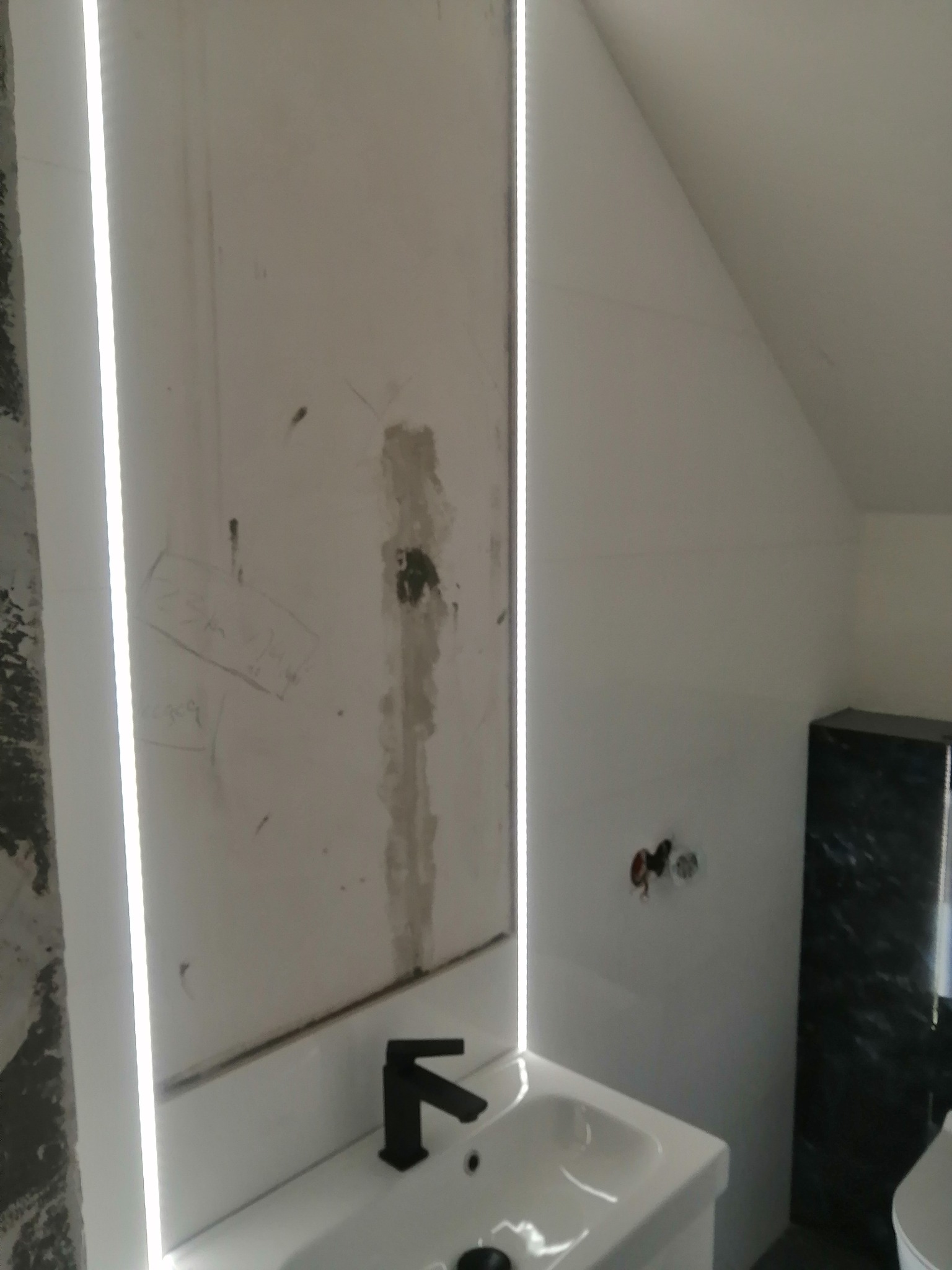 Łazienka w trakcie remontu: ściana z fragmentami starej glazury, nowa umywalka z czarną armaturą i podświetleniem LED, częściowo wykończona ściana z czarnym marmurem.