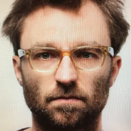 Portret mężczyzny z brodą i okularami w bursztynowych oprawkach na jasnym tle.