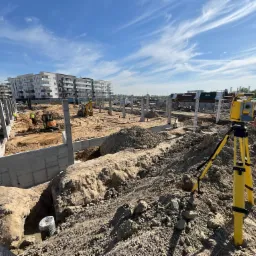 Żółty teodolit na trójnogu na placu budowy z widocznymi fundamentami, kolumnami, koparkami i budynkami mieszkalnymi w tle pod błękitnym niebem.