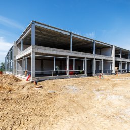 SOLIDGEO Sp. z o.o. - Rozległy plac budowy z dwupiętrowym budynkiem o betonowo-stalowej konstrukcji szkieletowej. Widoczne koparki JCB, czerwone rury instalacyjne i świeżo przekopana ziemia pod błękitnym niebem.