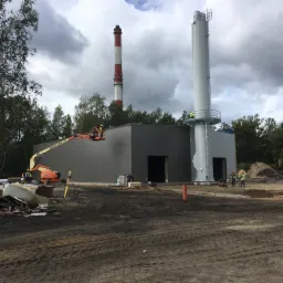 Budowa przemysłowa: montaż konstrukcji z dwoma kominami, w tle las, pracownicy w pomarańczowej platformie podnośnika teleskopowego i żółtych kamizelkach, ziemia rozjeżdżona przez maszyny.