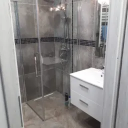 Nowoczesna łazienka z kabiną prysznicową typu walk-in, umywalką z szafką w kolorze białym, szarymi płytkami ściennymi z czarnymi akcentami i beżową podłogą z płytek imitujących kamień.